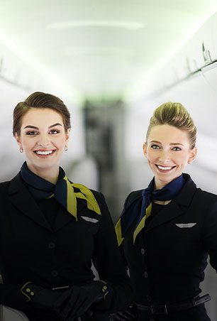 Duas comissárias de bordo posando para uma foto dentro de um avião, sorrindo e vestindo uniformes.