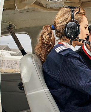 Um homem e uma mulher dentro de um pequeno avião, usando fones de ouvido, preparados para decolar.